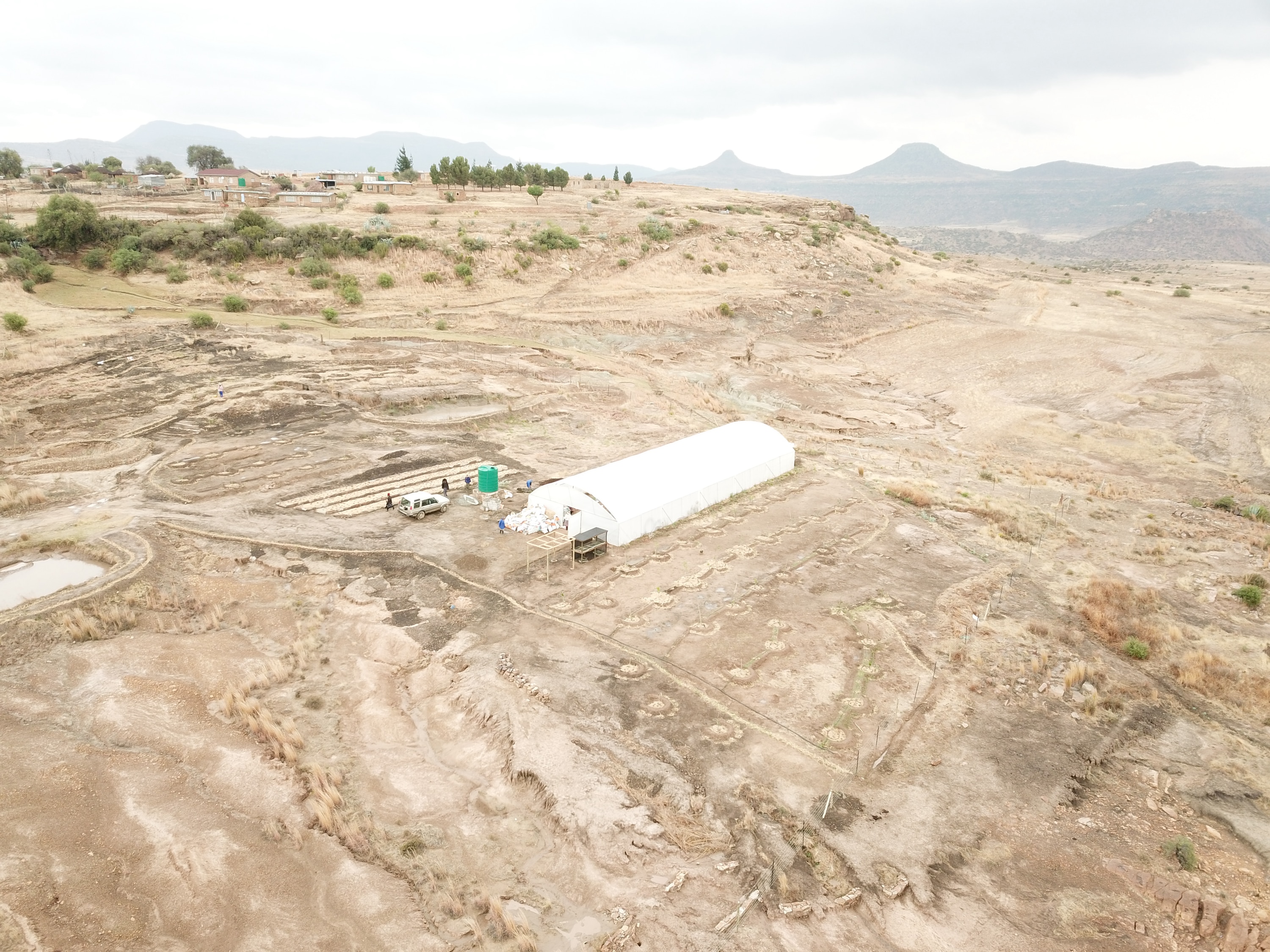 Oblique aerial view of the whole Morobong site: pale bare ground, the white polytunnel with a green water tank beside it, a block of small dark rectangles that are the raised beds, and a village and flat-topped mountains on the horizon