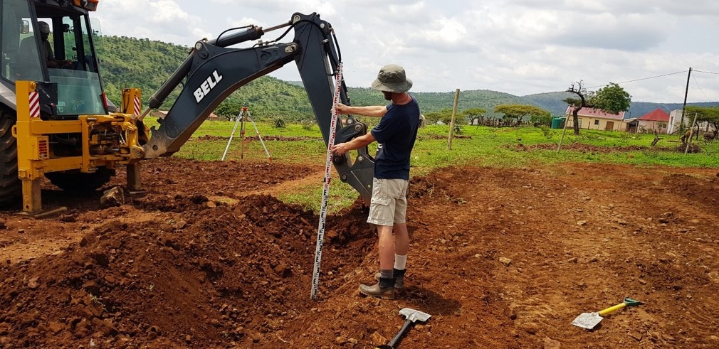 Rory Clark holding a surveying staff beside a Bell excavator, with a level on a tripod behind him